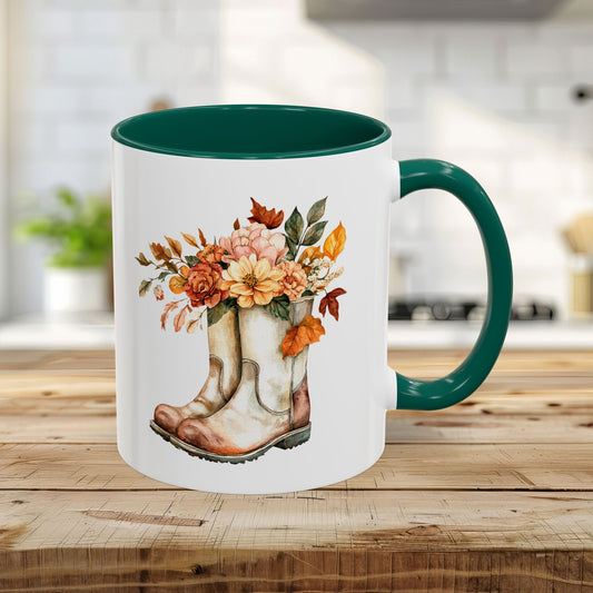 White 11oz ceramic mug with a green handle and interior, featuring a watercolor design of worn boots overflowing with autumn flowers.