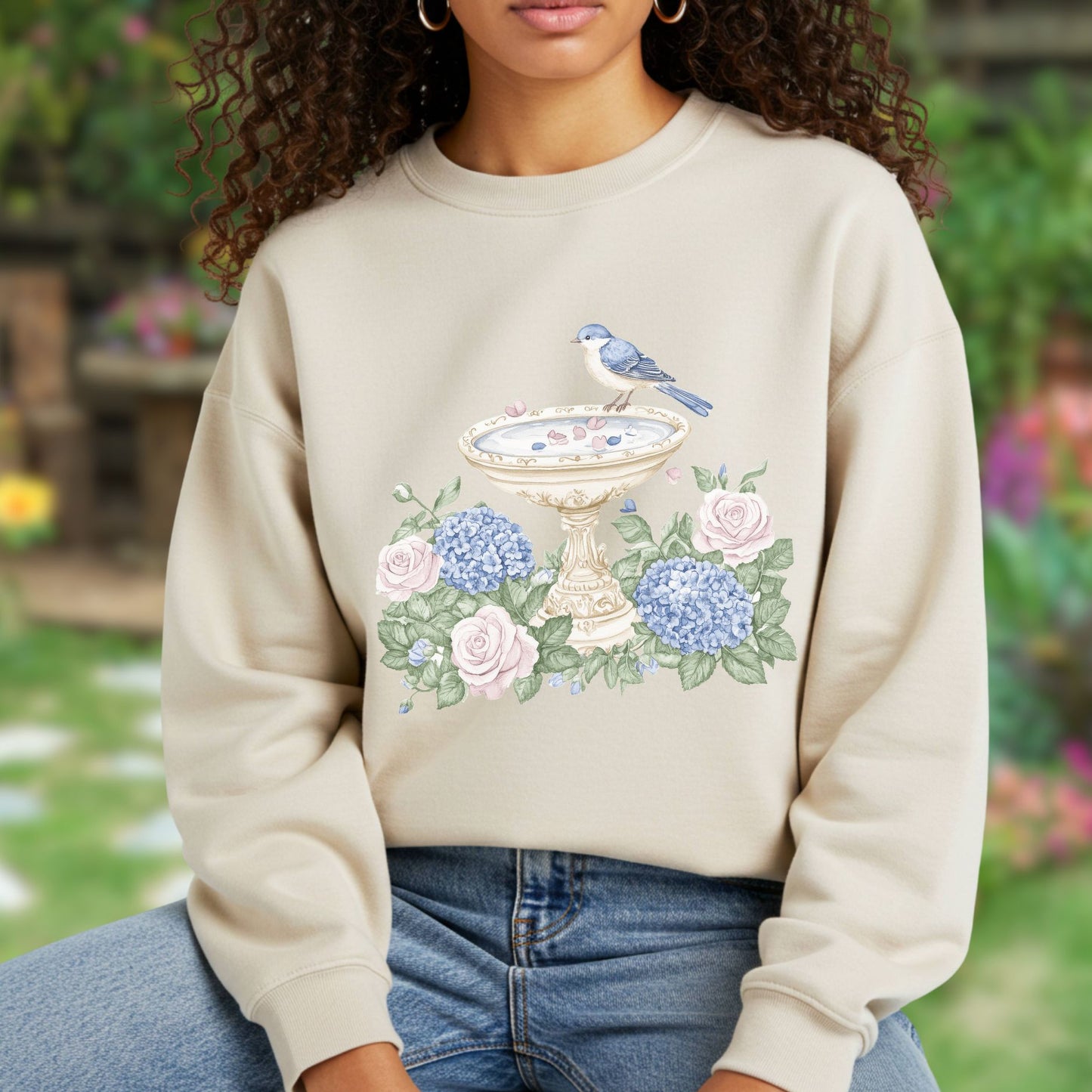 Beige sweatshirt with a watercolor bird bath, blue bird, pink roses, and blue hydrangeas design.