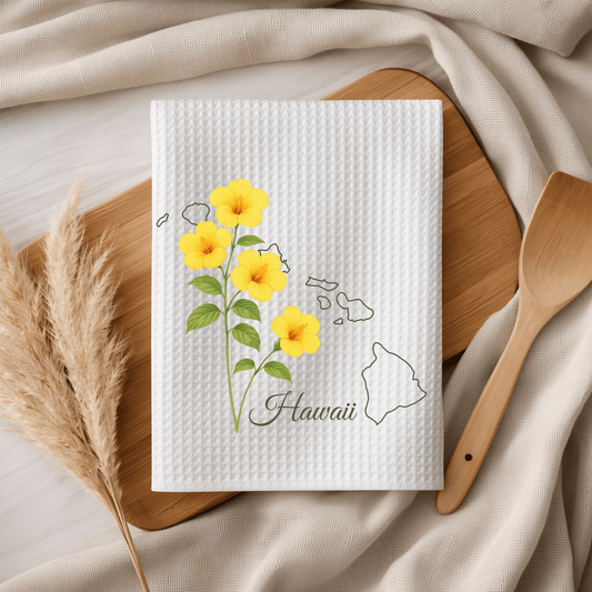 White microfiber tea towel with yellow hibiscus flowers, a Hawaii map outline, and "Hawaii" text on a wooden board.