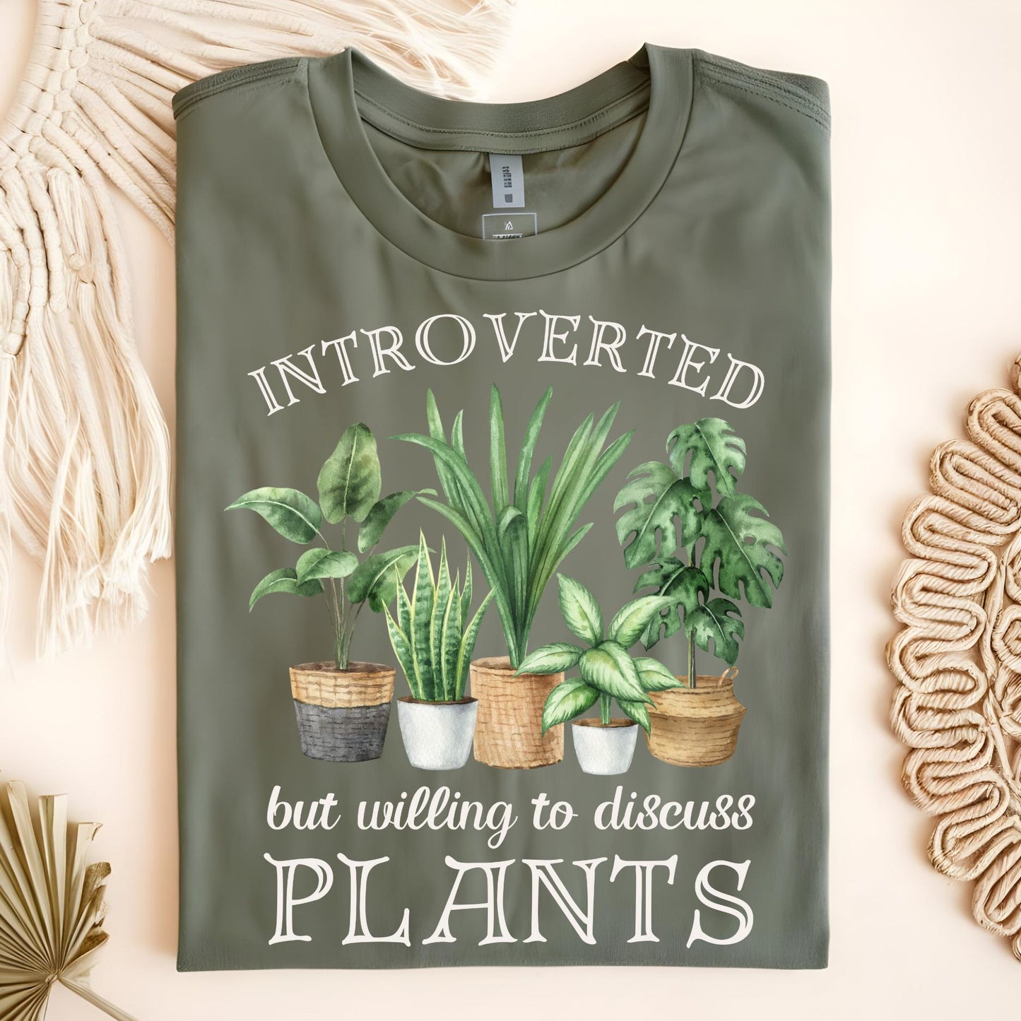 Folded green t-shirt with house plant design and text on a light background that reads "Introverted but willing to discuss plants"