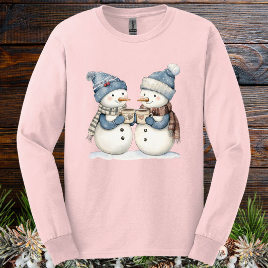 Light pink long sleeve tee with two snowpeople in blue hats, mittens, and scarves, holding hot chocolate.