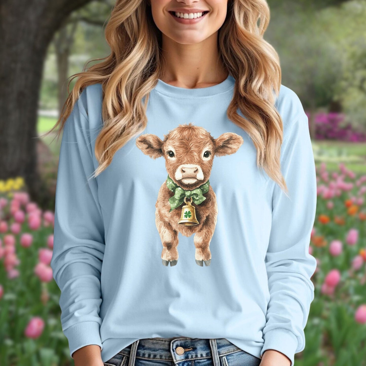 Light blue long-sleeve tee with a watercolor baby Highland cow wearing a green bow and clover bell.