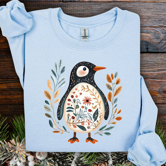 Light blue sweatshirt with folk art penguin, botanical patterns, orange beak, and striped legs.