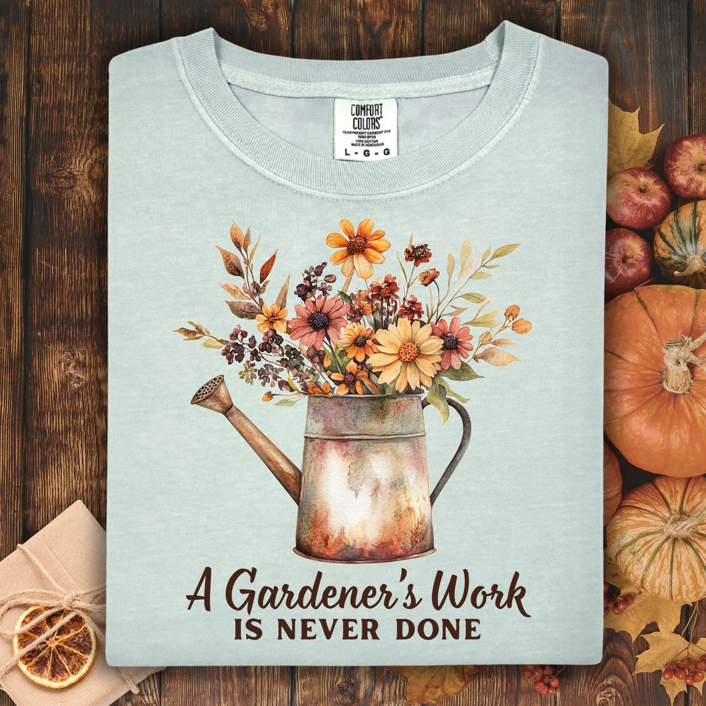 Bay blue Comfort Colors t-shirt with a watering can full of fall flowers and "A Gardener's Work Is Never Done" text.