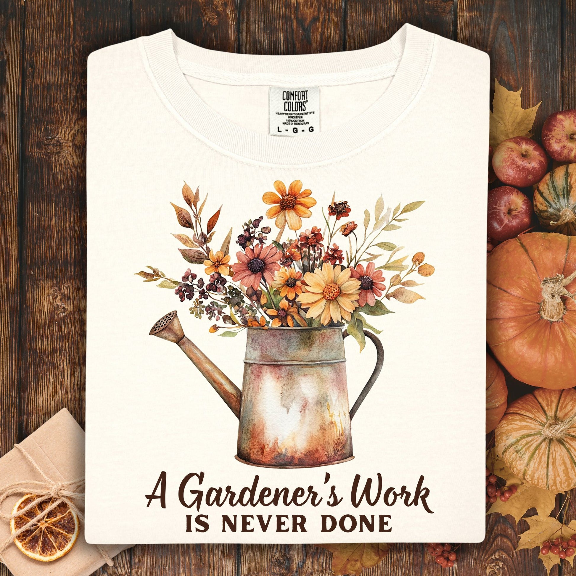 Ivory Comfort Colors tee with a watercolor watering can full of autumn flowers and "A Gardener's Work Is Never Done" text.