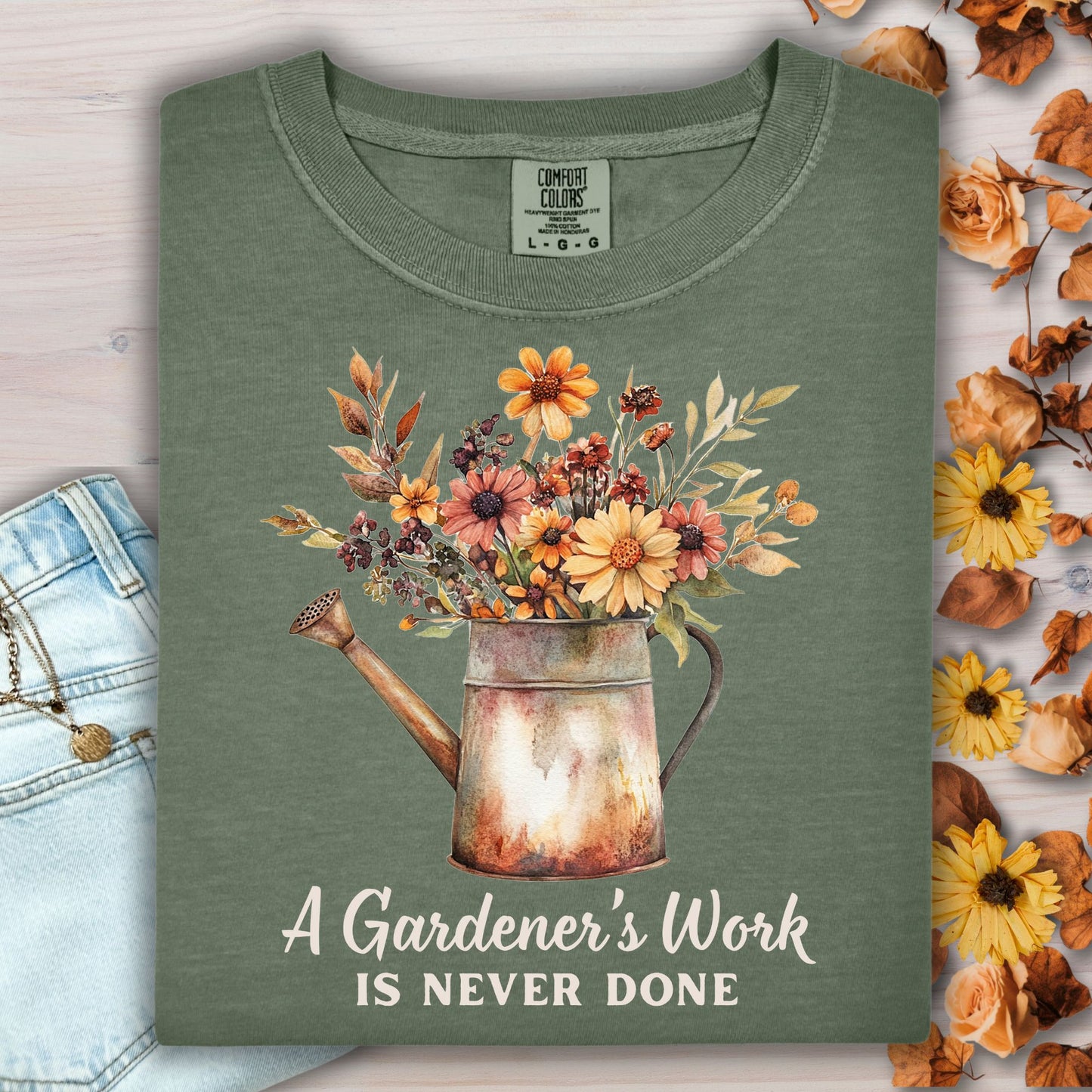Moss green Comfort Colors t-shirt with a watering can full of autumn flowers and "A Gardener's Work Is Never Done" text.