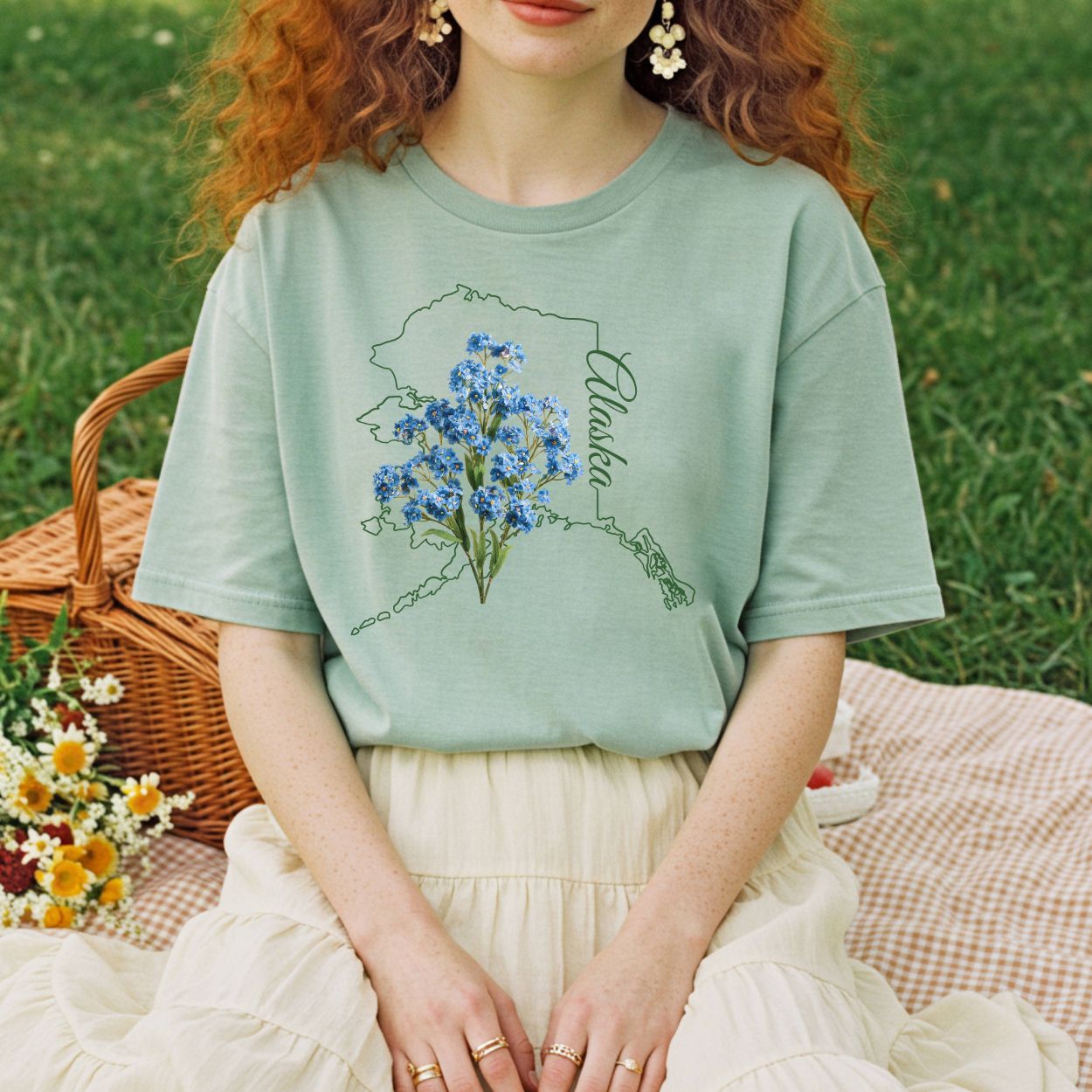 Relaxed-fit, garment-dyed sage green t-shirt with an Alaska map outline and blue forget-me-not flowers.