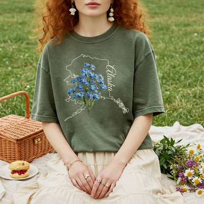 Garment-dyed olive green t-shirt with an Alaska outline and blue forget-me-not flowers.