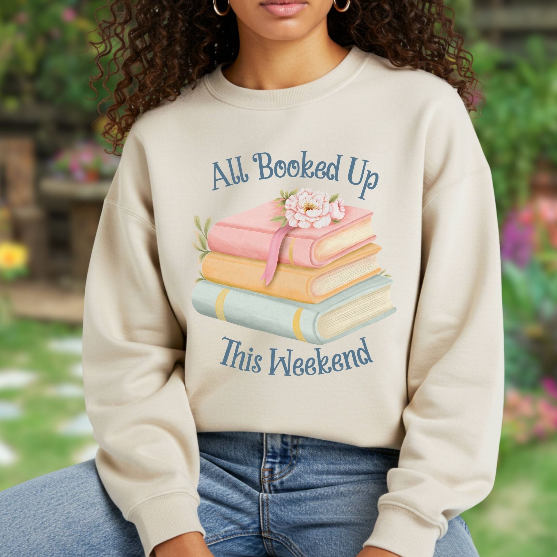 Woman wearing a heathered beige crewneck sweatshirt with a watercolor stack of books and "All Booked Up This Weekend" text.