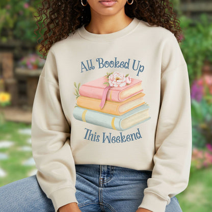 Woman wearing a heathered beige crewneck sweatshirt with a watercolor stack of books and "All Booked Up This Weekend" text.