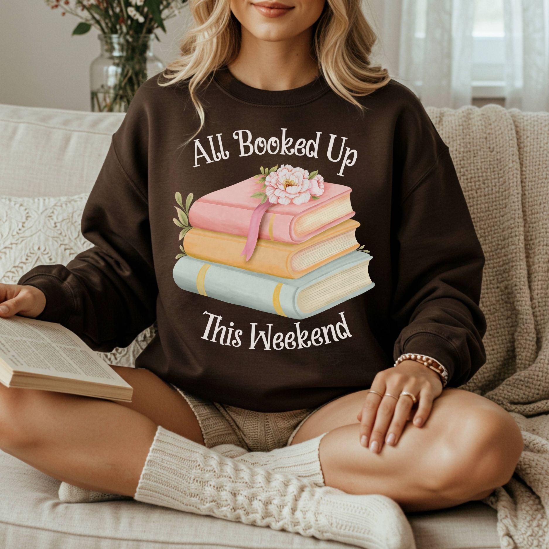 Woman in a brown "All Booked Up This Weekend" sweatshirt with watercolor books, reading on a sofa.