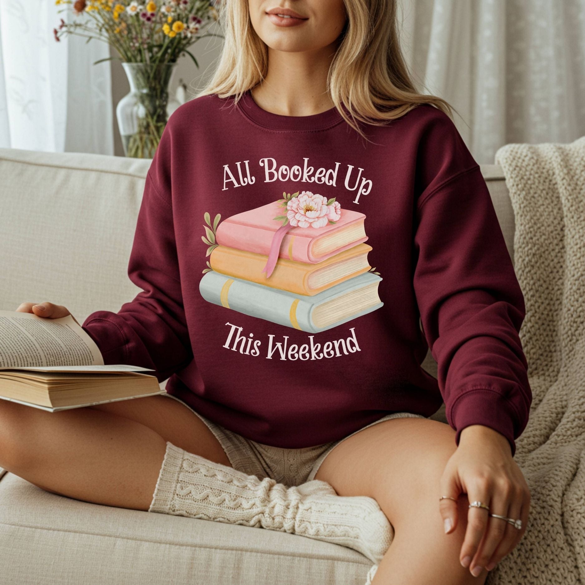 Woman in a maroon crewneck sweatshirt with "All Booked Up This Weekend" and a watercolor stack of books.