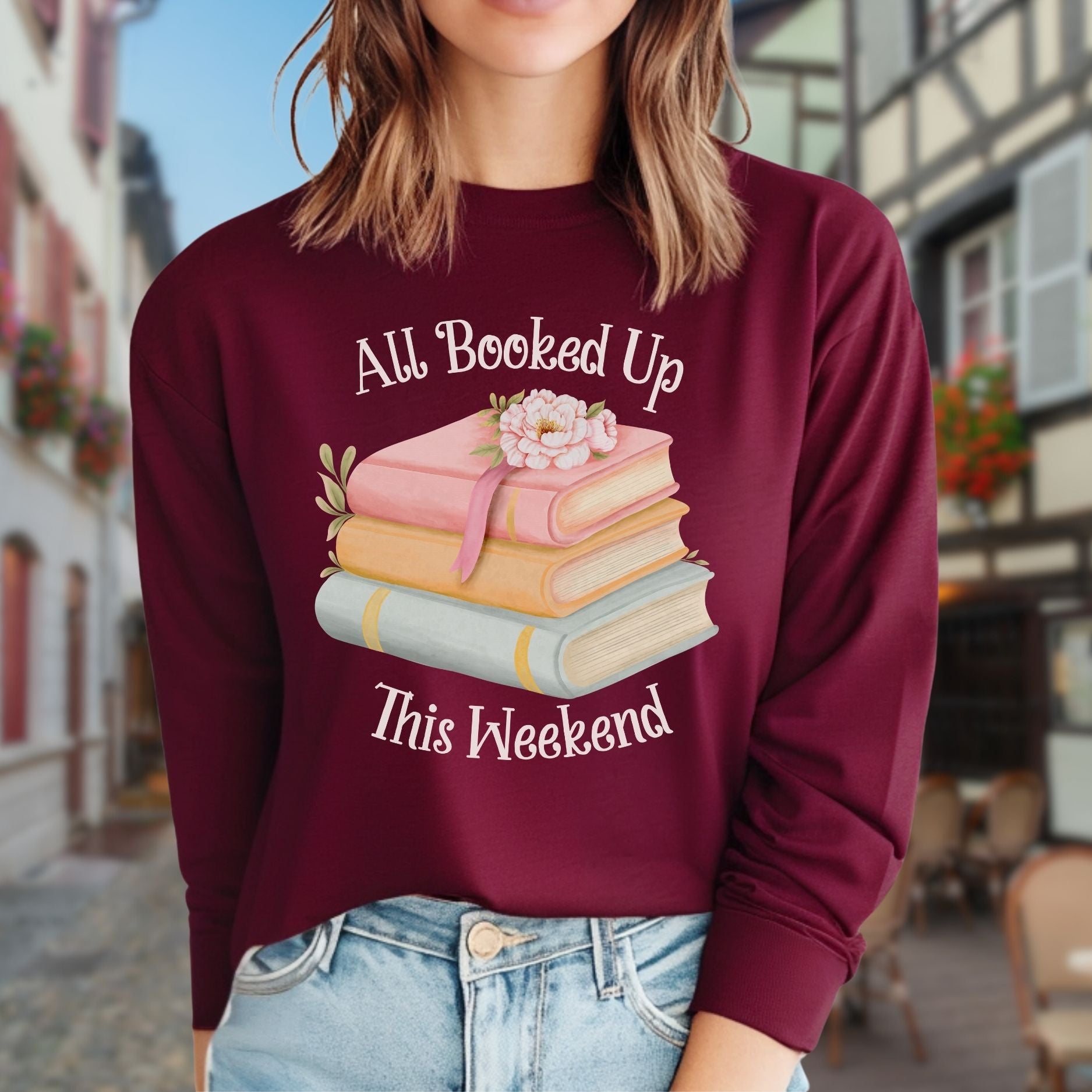 Maroon long-sleeve tee with "All Booked Up This Weekend" text, pastel books, and a floral illustration.