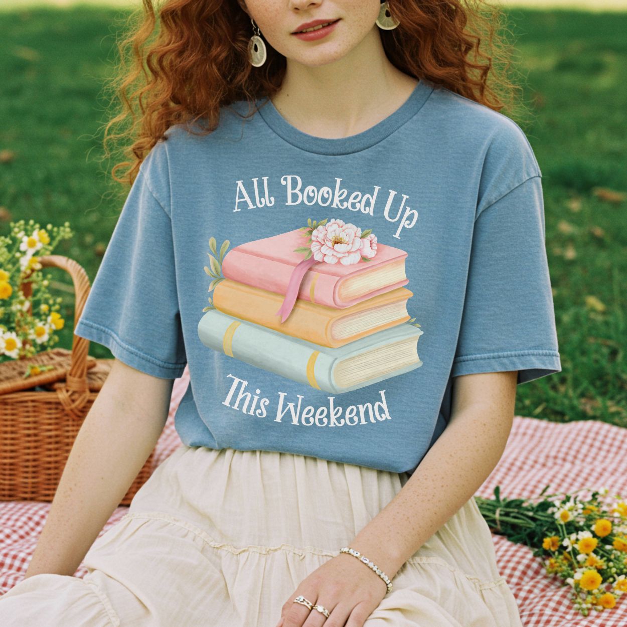 Garment-dyed blue t-shirt with "All Booked Up This Weekend" text and pastel book stack design.