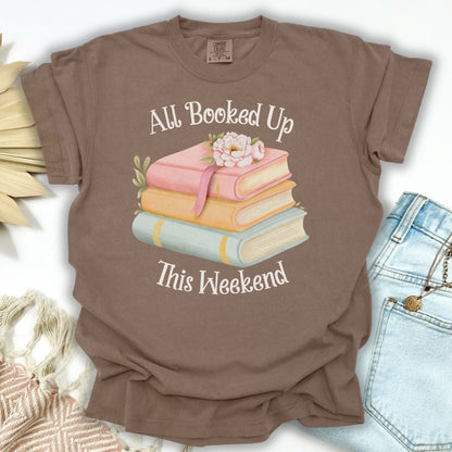 Espresso garment-dyed t-shirt with "All Booked Up This Weekend" text and a stack of pastel books with flowers.
