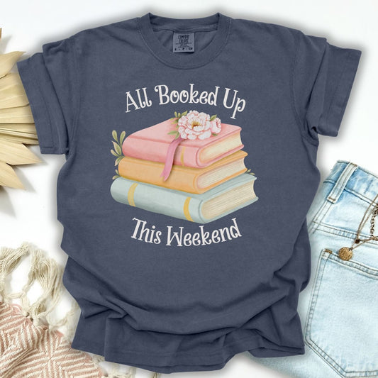 Denim blue garment-dyed t-shirt with "All Booked Up This Weekend" text and pastel book stack design.