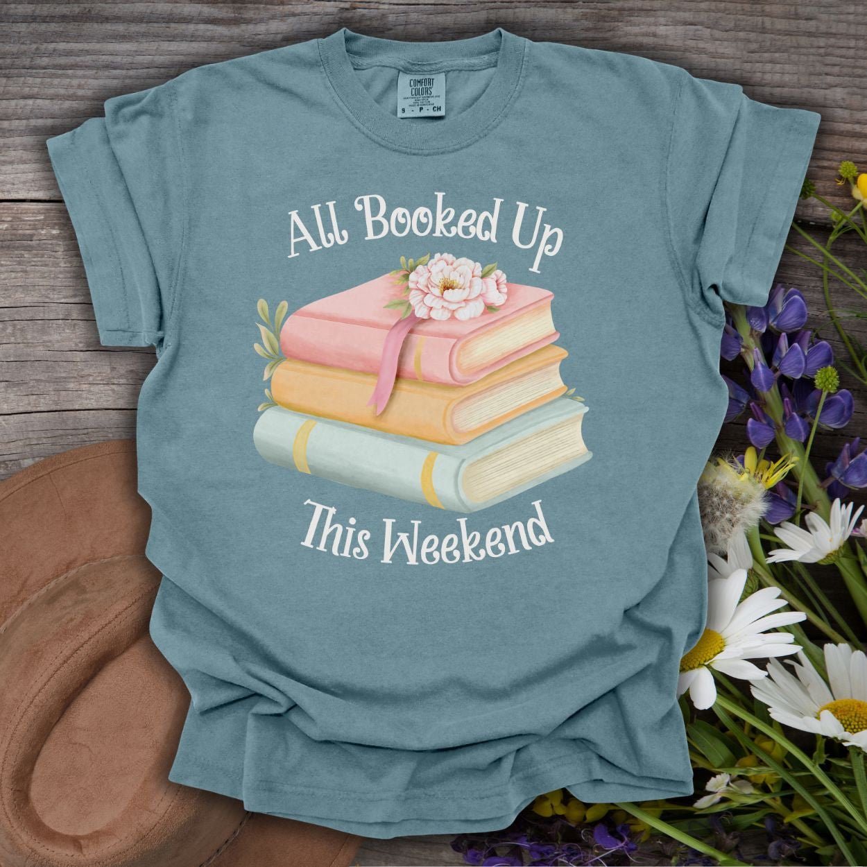 Ice Blue garment-dyed cotton t-shirt with "All Booked Up This Weekend" text and pastel book stack graphic.
