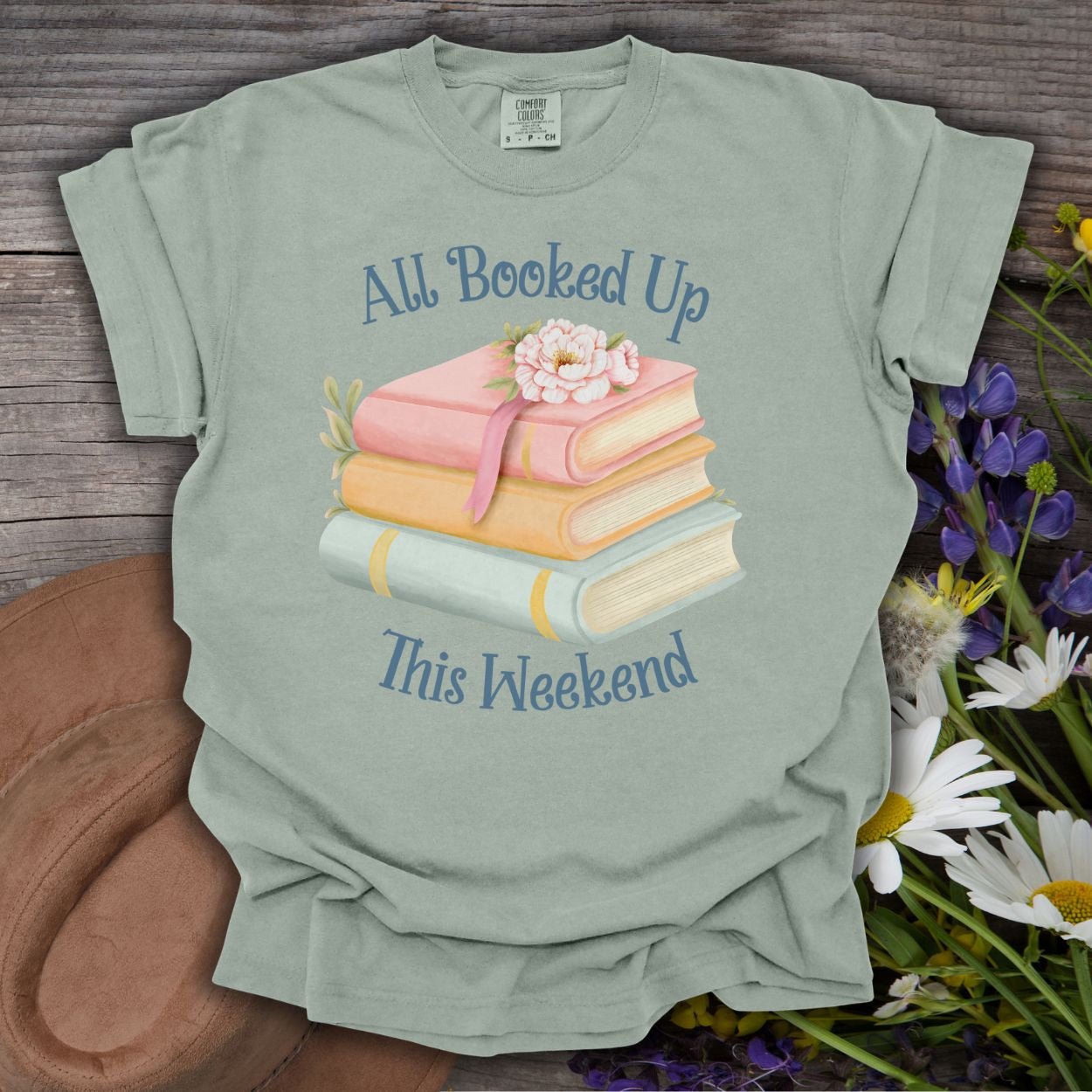 Bay garment-dyed cotton t-shirt with "All Booked Up This Weekend" and a stack of pastel books with flowers.