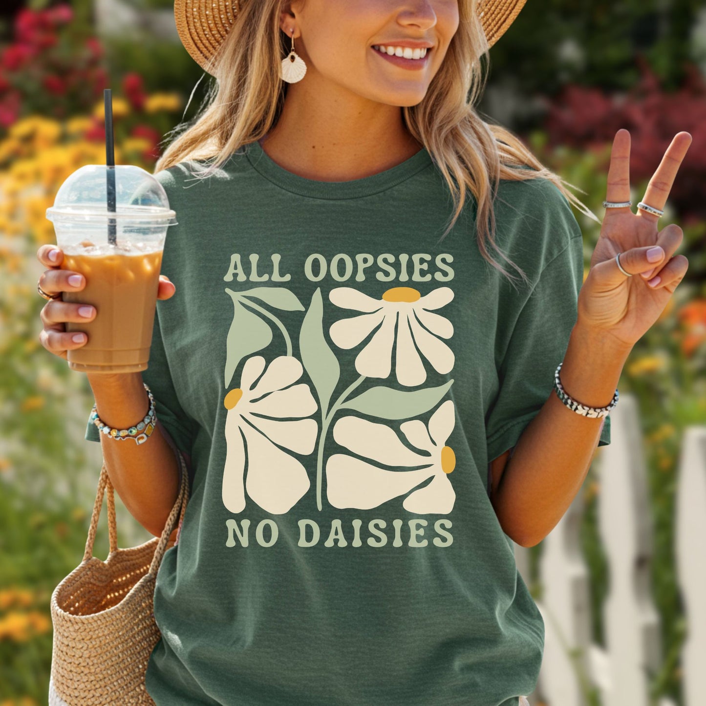 Woman in a forest green "All Oopsies No Daisies" retro floral t-shirt, straw hat, and earrings, holding iced coffee.