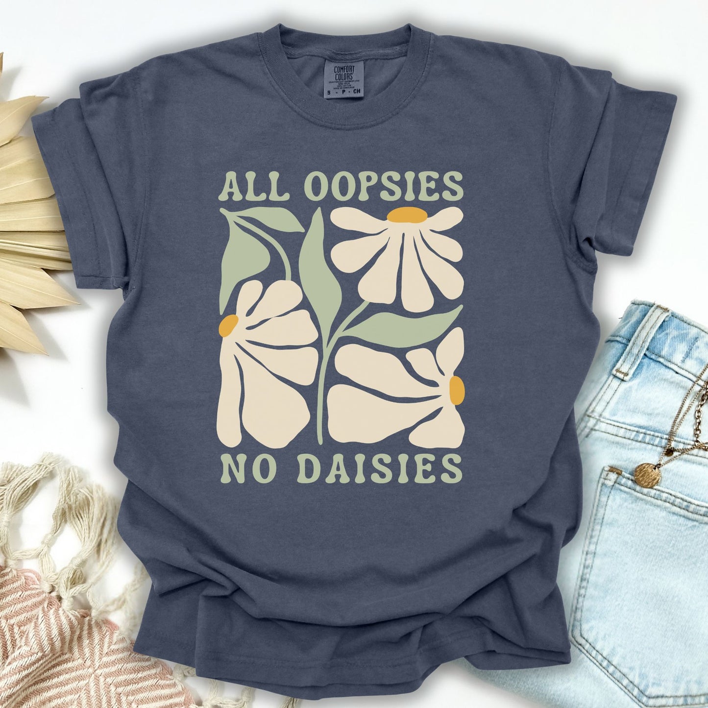Denim blue garment-dyed cotton t-shirt with "All Oopsies No Daisies" text and retro floral design.