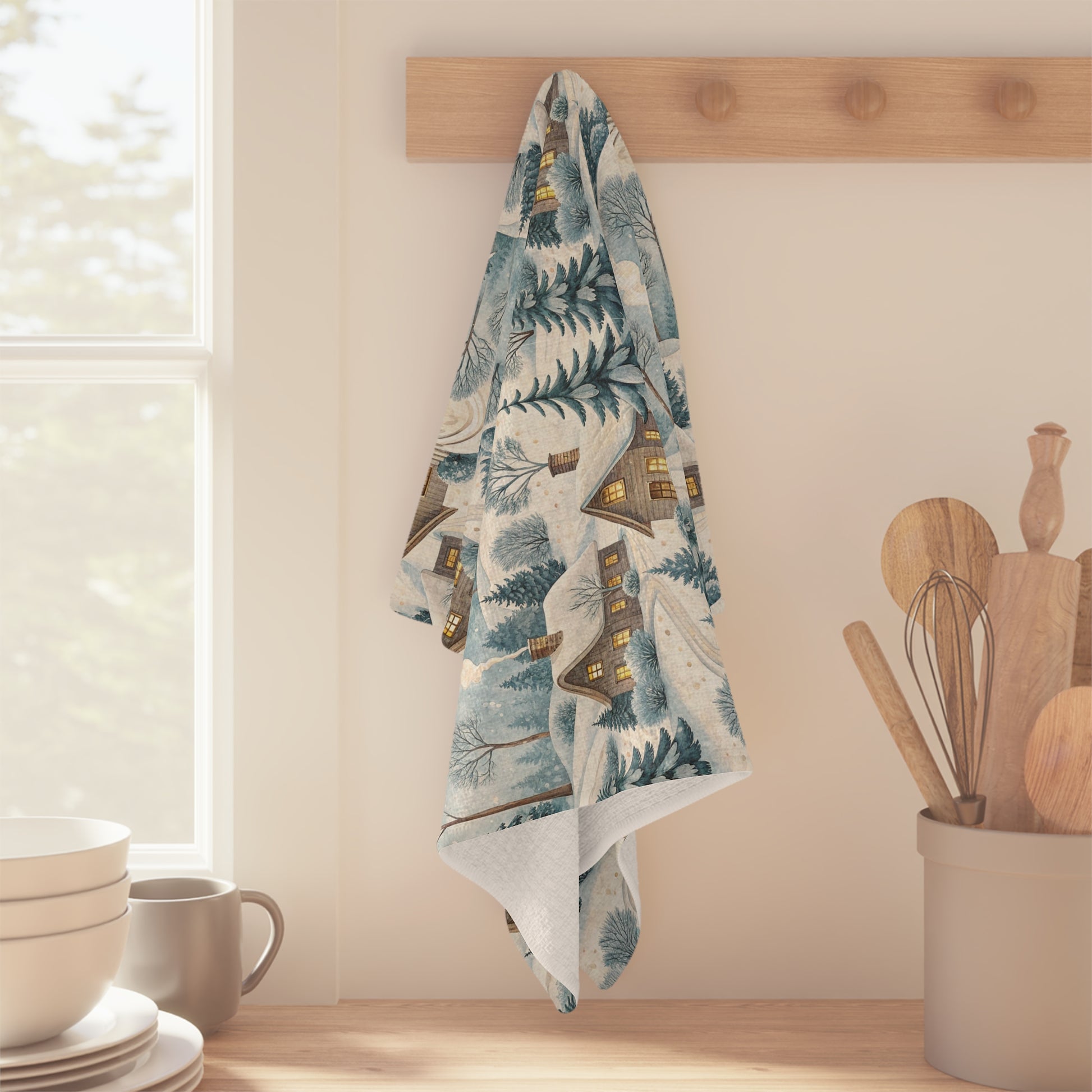 Microfiber tea towel with a muted blue and ochre Alpine winter village design, hanging on a wooden hook.
