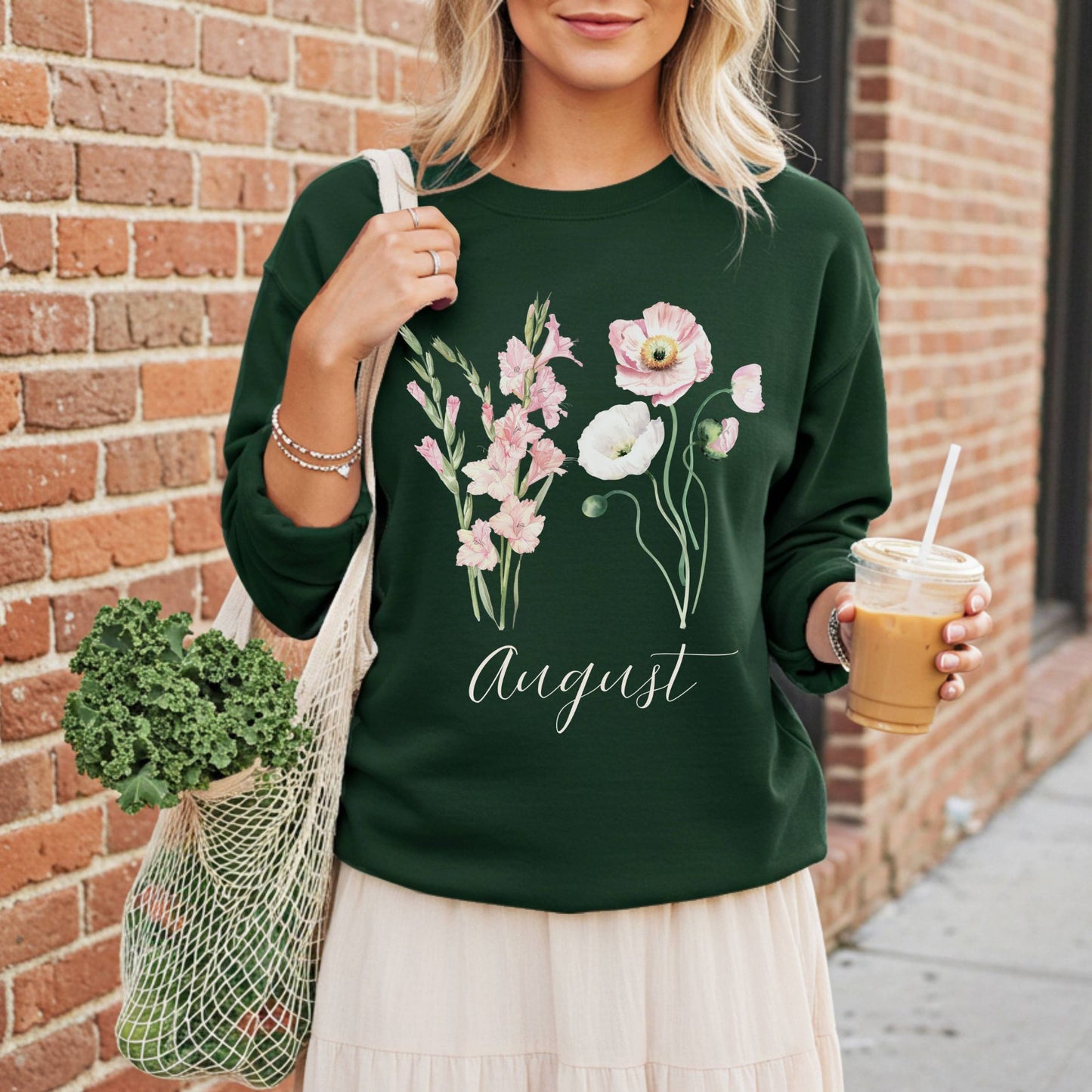 Dark green sweatshirt with watercolor gladiolus and poppy flowers, "August" script, worn by a person holding a coffee and market bag.