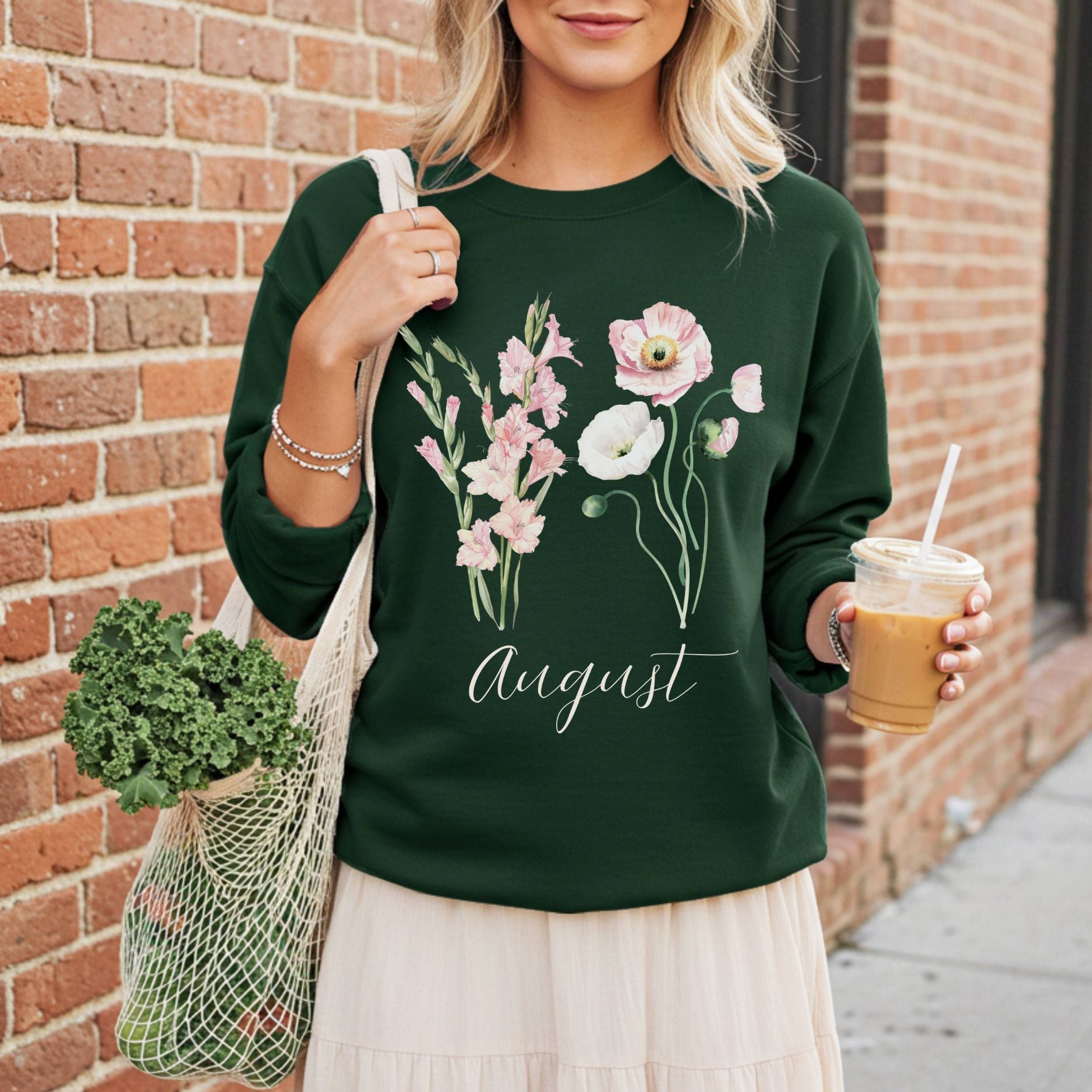 Dark green sweatshirt with watercolor gladiolus and poppy flowers, "August" script, worn by a person holding a coffee and market bag.