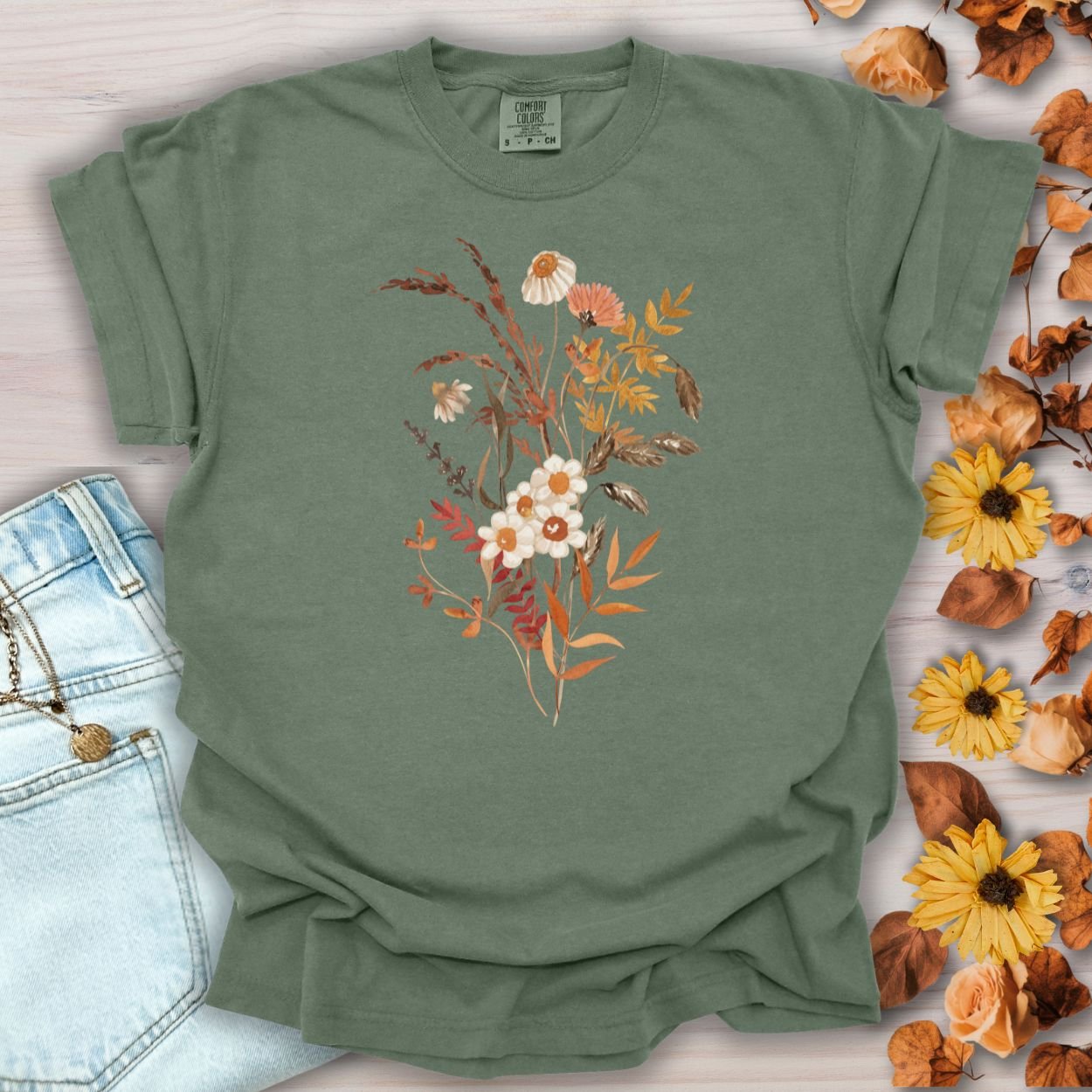 Moss green S-size Autumn Botanical T-shirt with a warm, nature-inspired bouquet print, next to jeans and dried leaves.