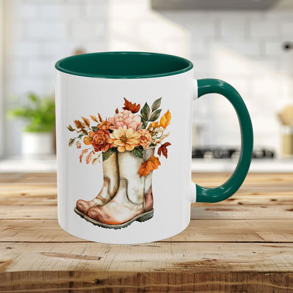 White 11oz ceramic mug with a green handle and interior, featuring a watercolor design of worn boots overflowing with autumn flowers.