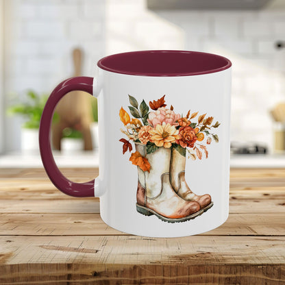 White ceramic mug with a maroon handle and interior, featuring a watercolor design of worn cowboy boots overflowing with autumn flowers and leaves.