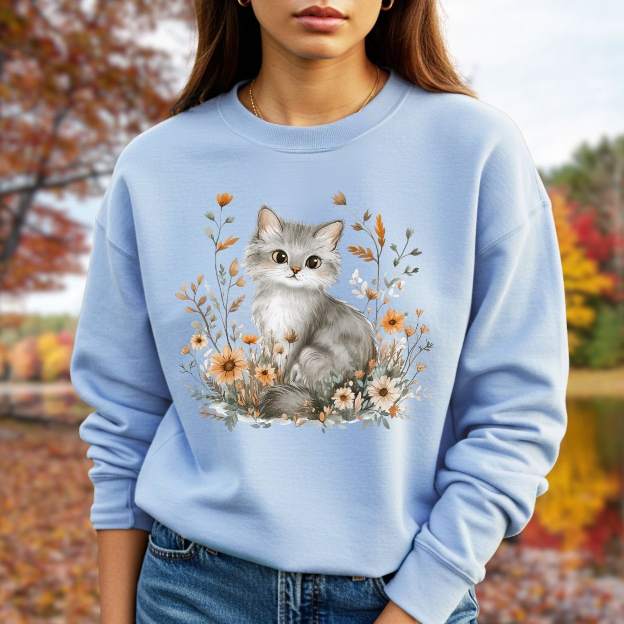 Light blue crewneck sweatshirt with a grey kitten among autumn wildflowers, worn by a woman outdoors.