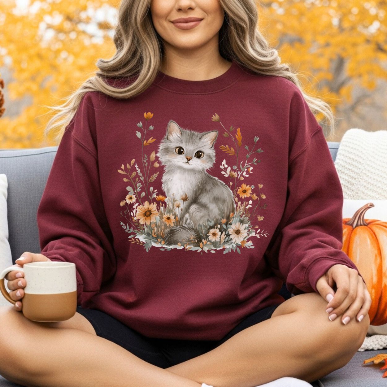 Woman in a maroon crewneck sweatshirt with a grey kitten and autumn wildflowers design, holding a mug.