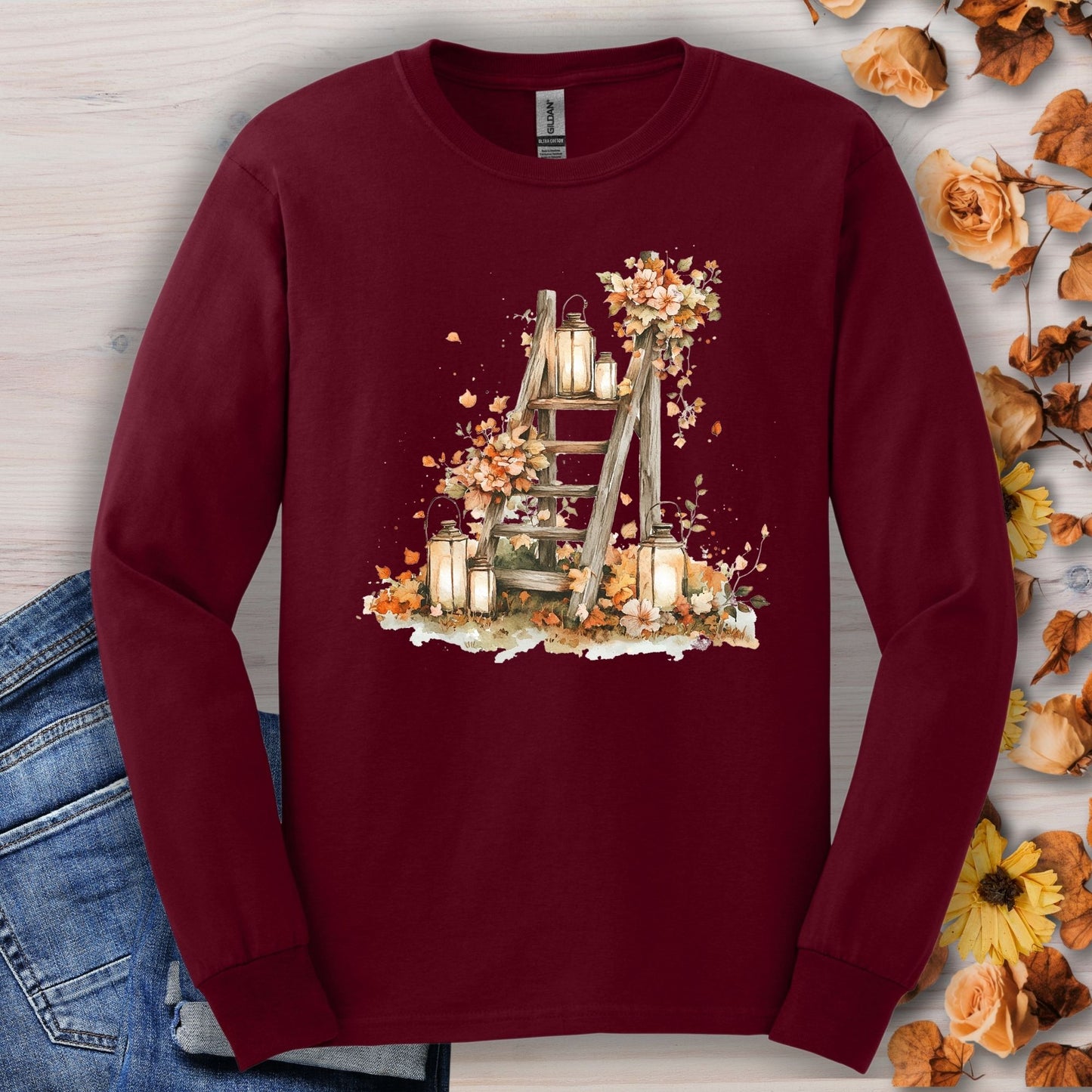 Maroon long sleeve tee with watercolor autumn lanterns, flowers, and leaves on a rustic ladder.