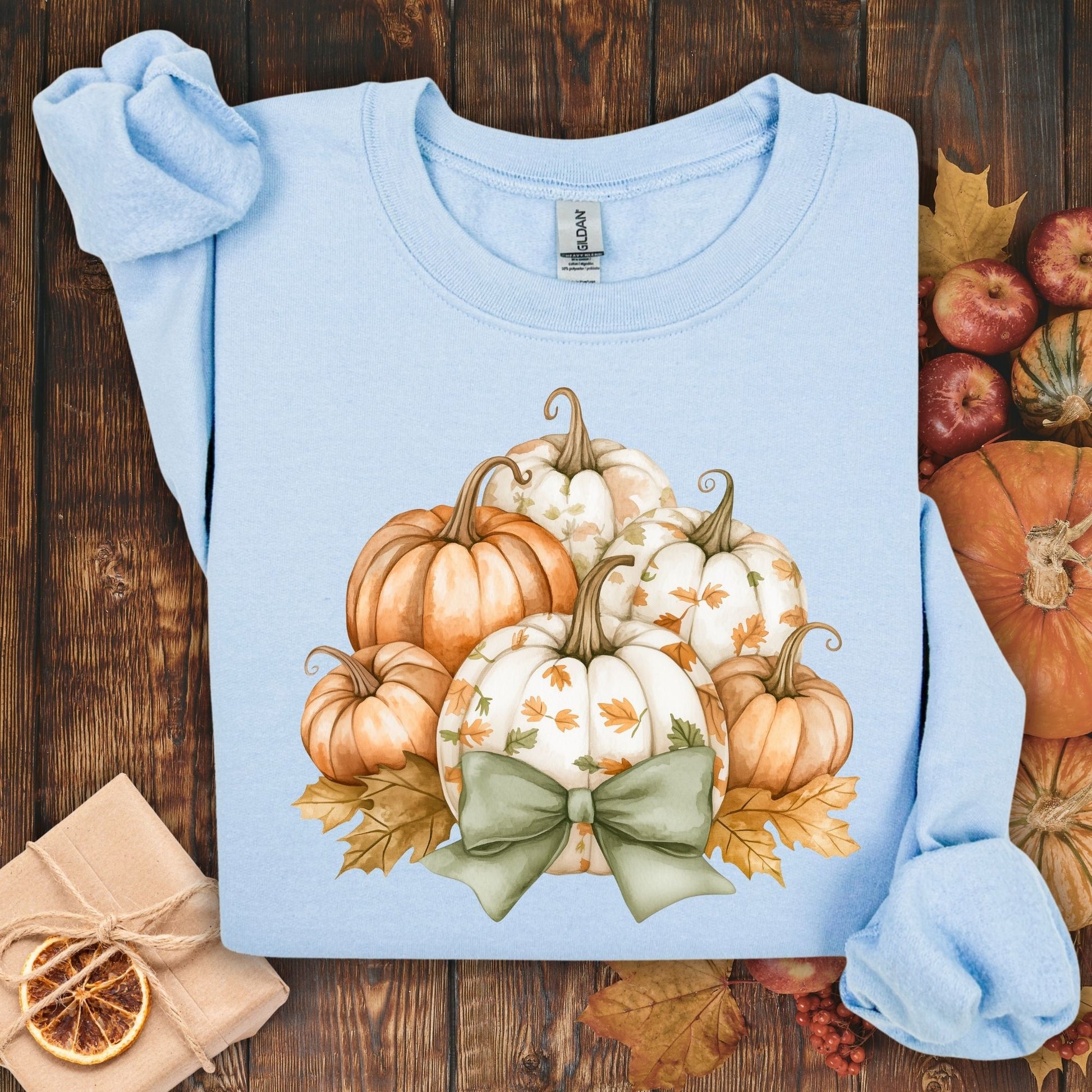 Light blue Gildan sweatshirt with a cottagecore pumpkin and green bow design, surrounded by fall leaves.