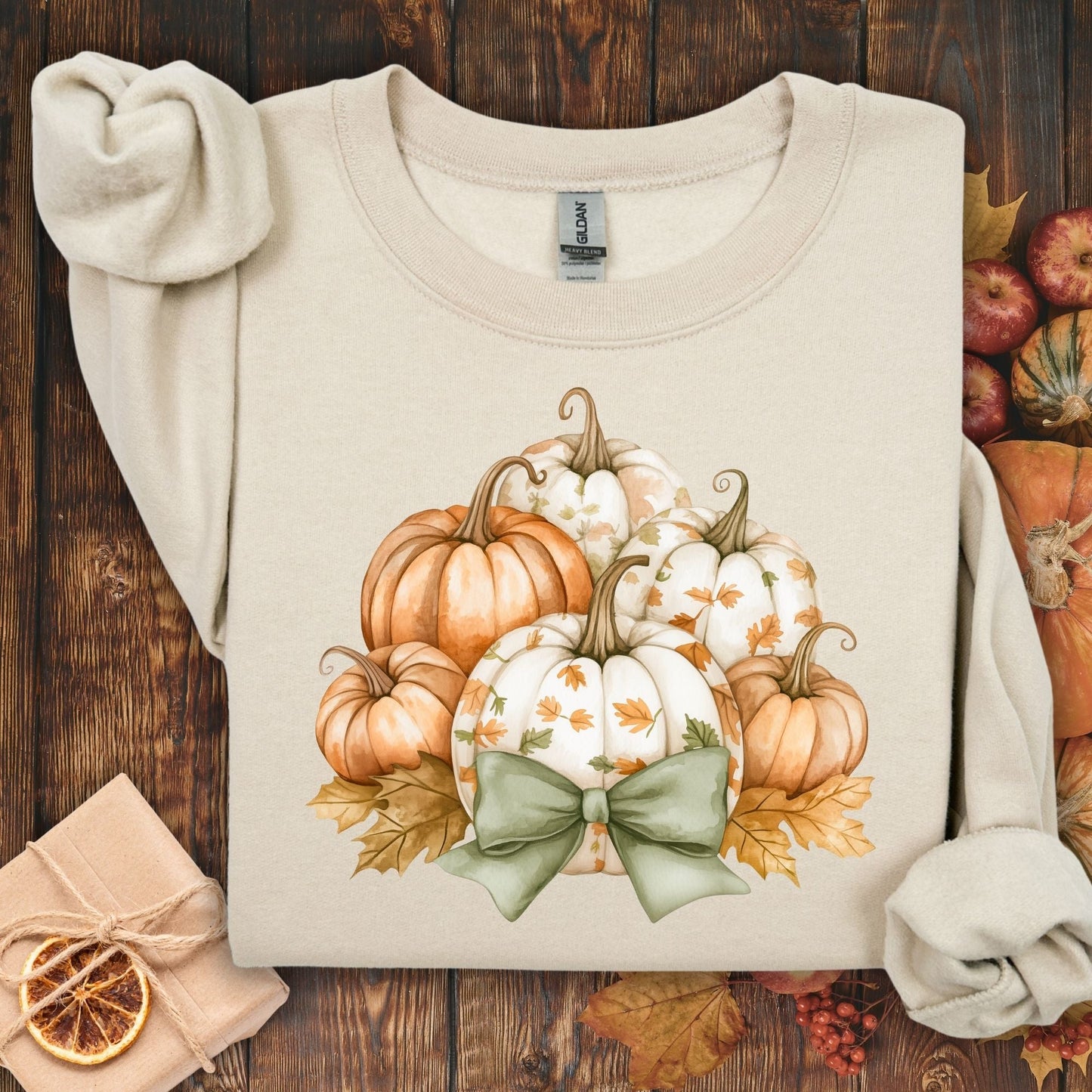 Sand Gildan sweatshirt with a cottagecore pumpkin and green bow design, surrounded by fall leaves.