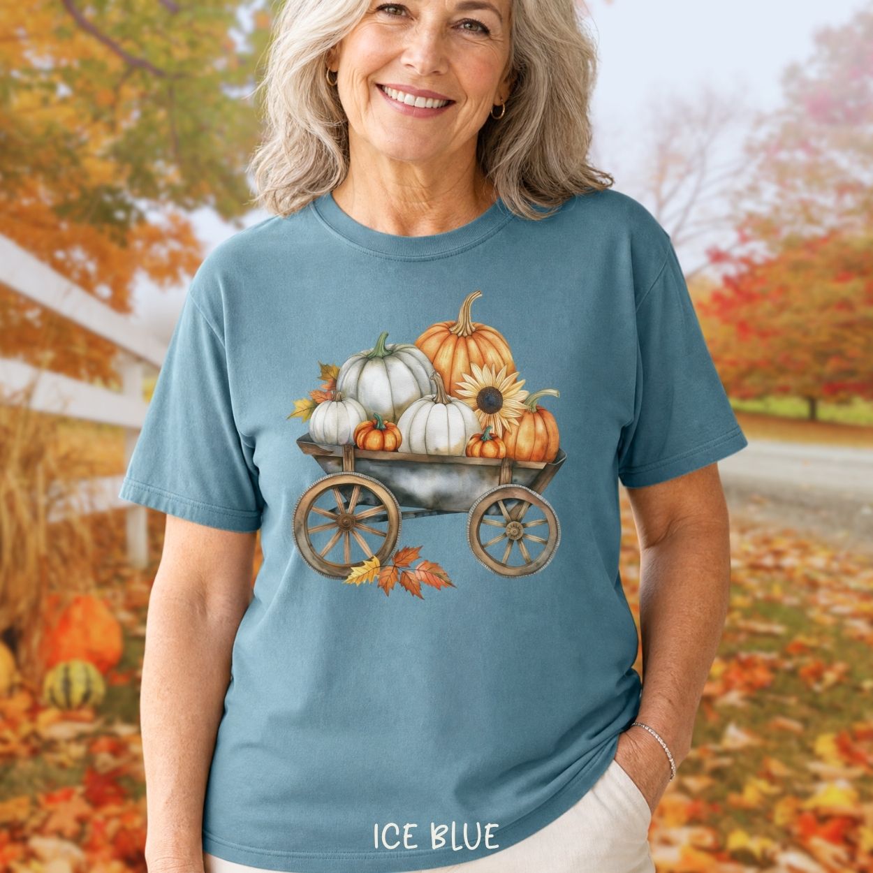 Woman in an ice blue t-shirt with a watercolor rustic wagon filled with white and orange pumpkins and sunflowers.
