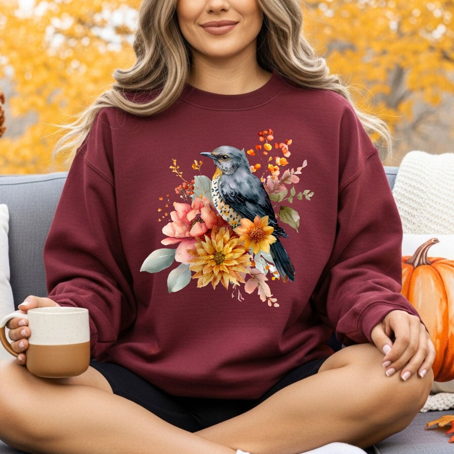 Maroon crewneck sweatshirt with a painterly bluebird and autumn floral design, worn by a woman outdoors.