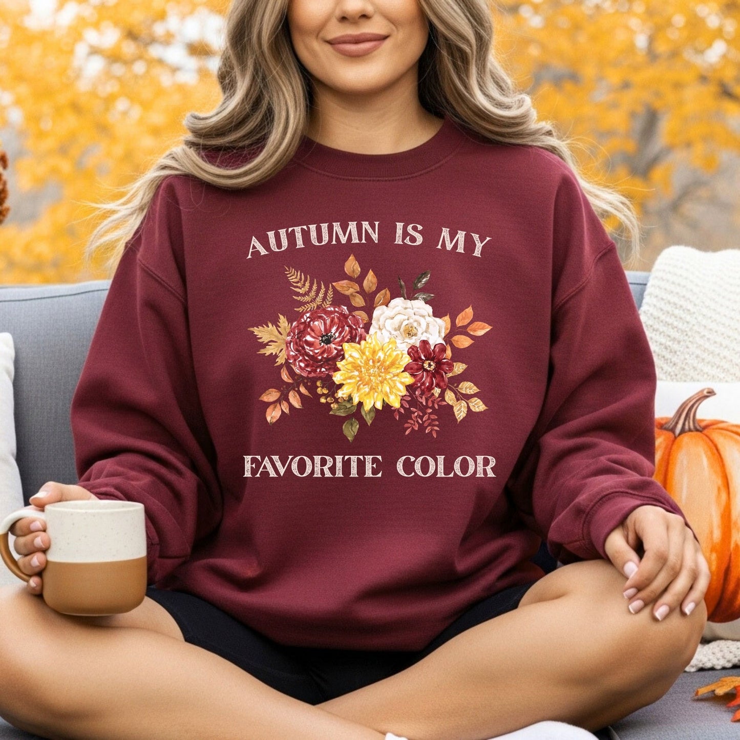 Woman wearing a maroon "Autumn Is My Favorite Color" sweatshirt with watercolor fall flowers, holding a mug.
