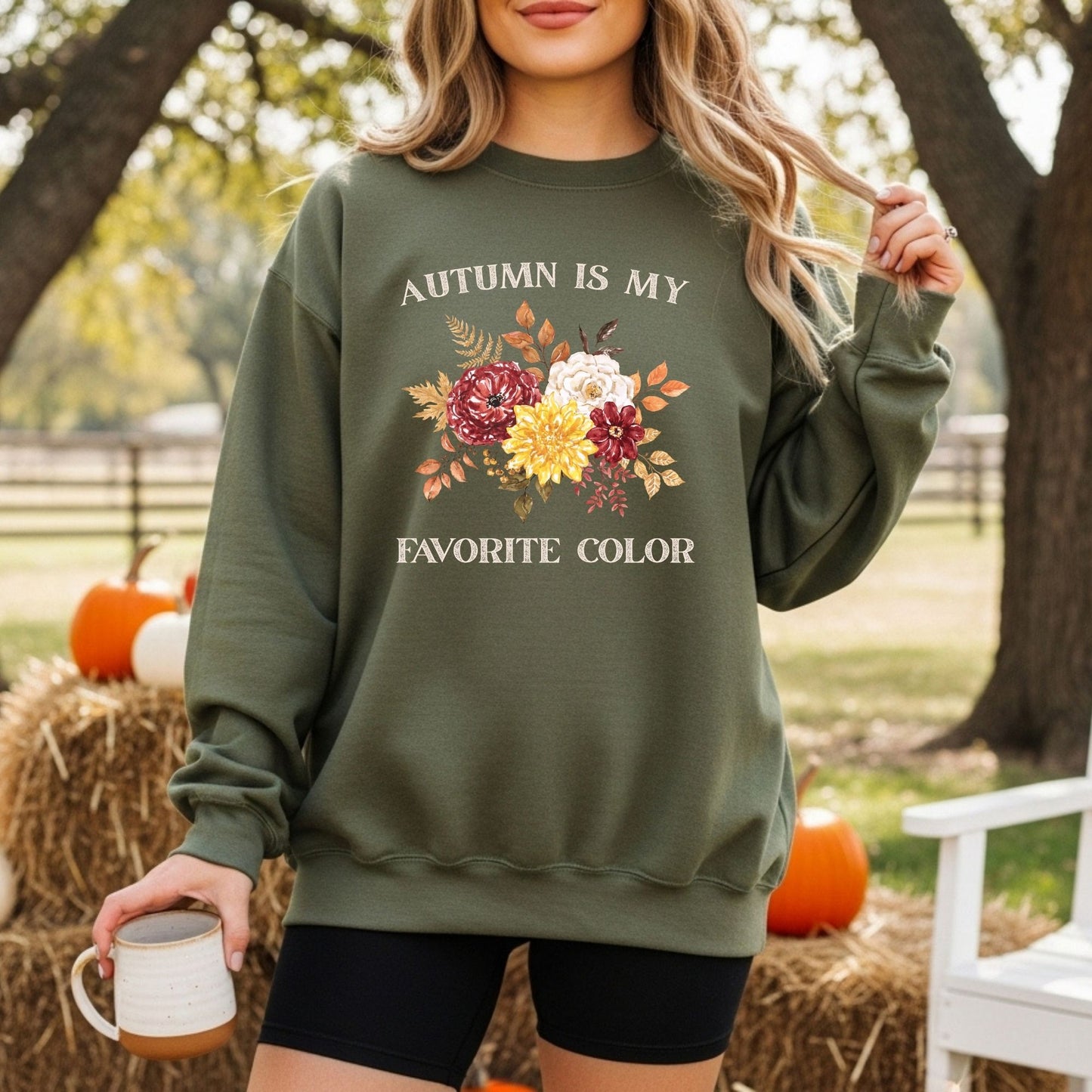 Olive green sweatshirt with "Autumn is My Favorite Color" text and watercolor fall flowers, worn by a woman outdoors.
