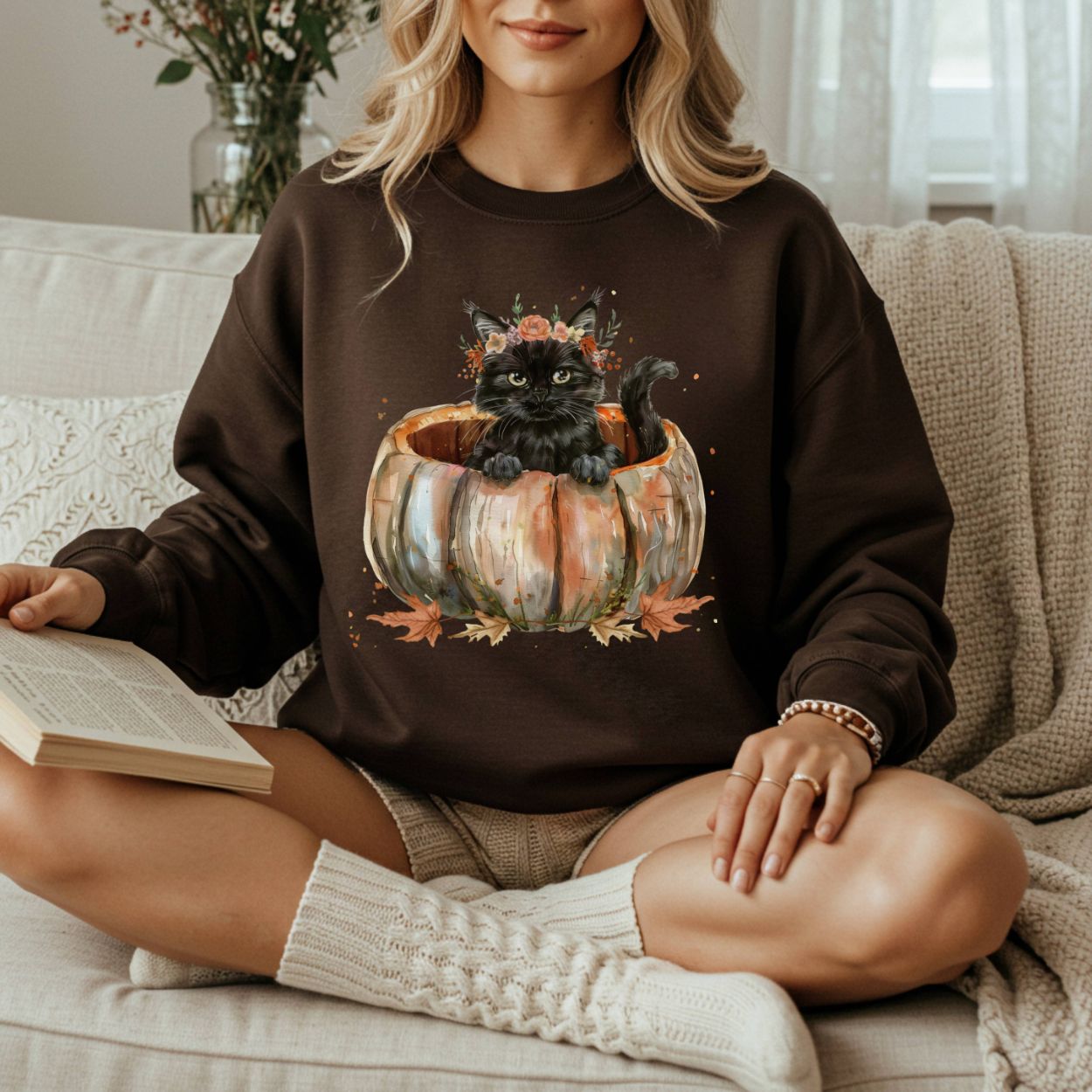 Woman in a brown sweatshirt with a watercolor black cat in a pumpkin and autumn flowers design.