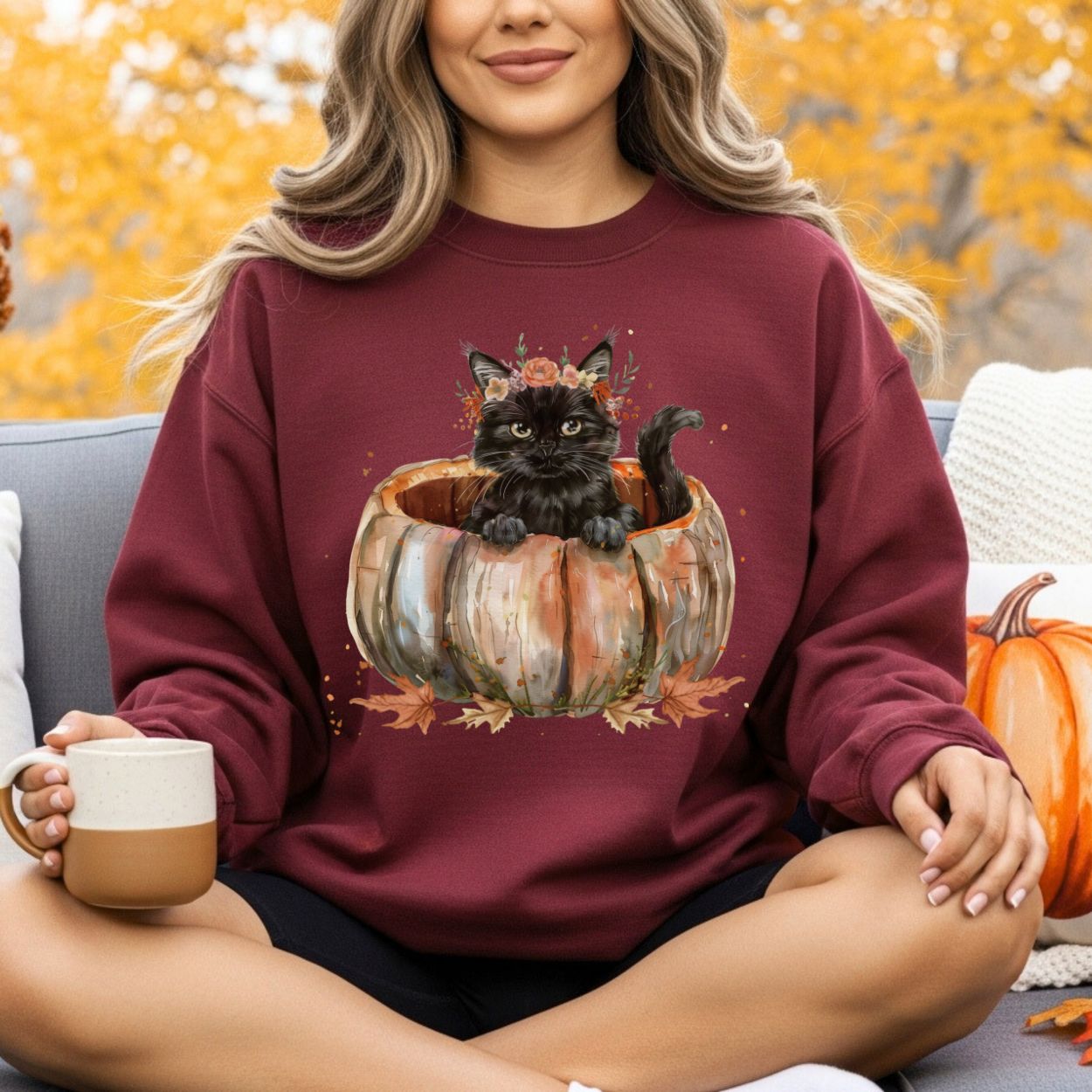 Woman in a maroon sweatshirt with a watercolor black cat in a pumpkin and autumn flowers.