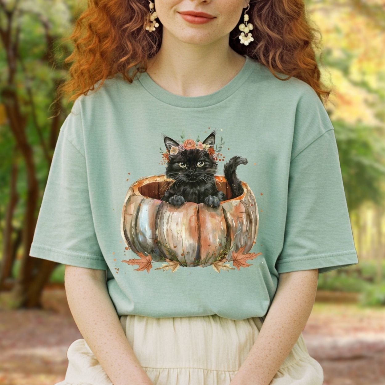 Woman wearing a sage green Comfort Colors t-shirt with a watercolor black cat in a pumpkin and fall flowers.