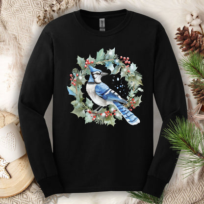 Black long sleeve tee with a watercolor blue jay in a holly wreath design, on a cozy winter background.