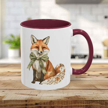 11oz ceramic mug with a watercolor fox wearing a green bow, maroon handle and interior, on a wooden surface.