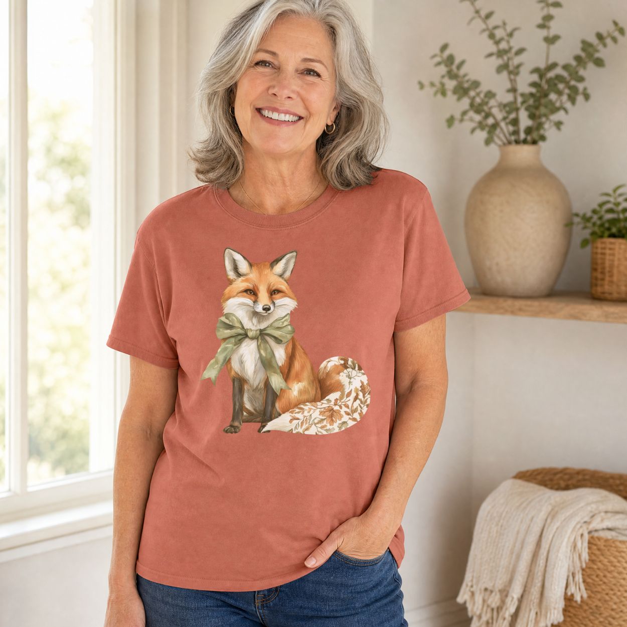 Woman in a terracotta garment-dyed t-shirt featuring a watercolor fox with a sage-green bow and leaf-patterned tail.