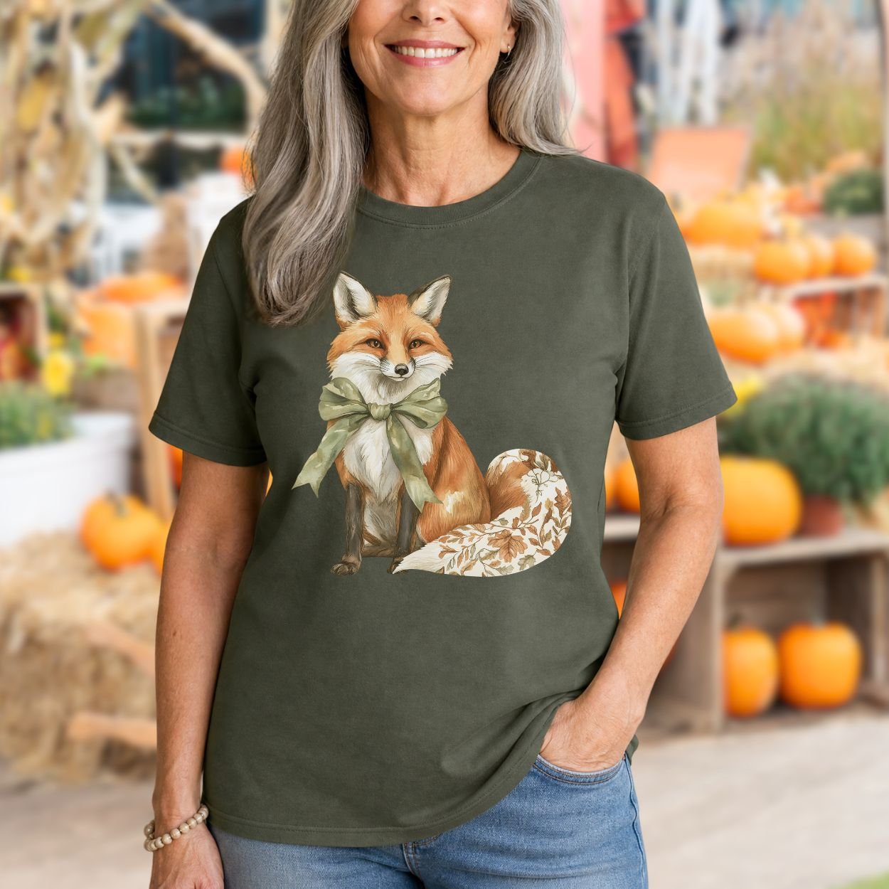 Woman in a relaxed-fit, garment-dyed olive green t-shirt featuring a watercolor fox with a sage green bow and leaf-patterned tail.
