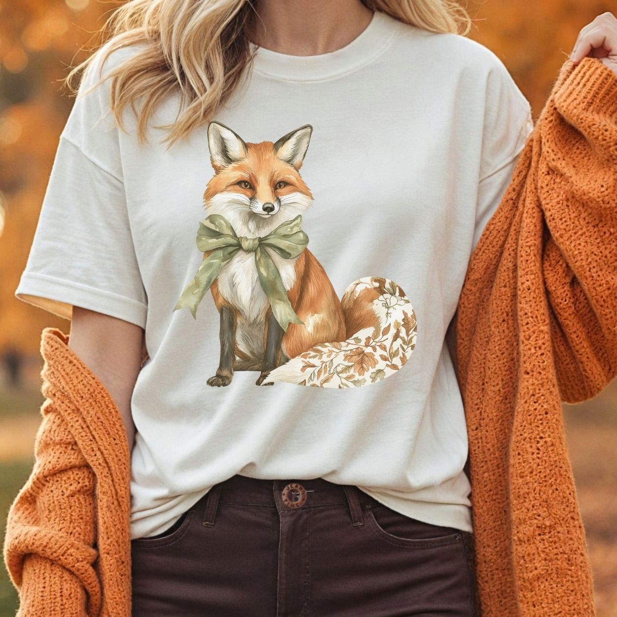 Garment-dyed cream t-shirt with a watercolor fox wearing a sage-green bow and a leaf-patterned tail.