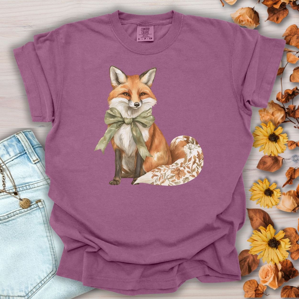 Berry S t-shirt with a watercolor fox, sage green bow, and leaf-patterned tail, on a rustic background.
