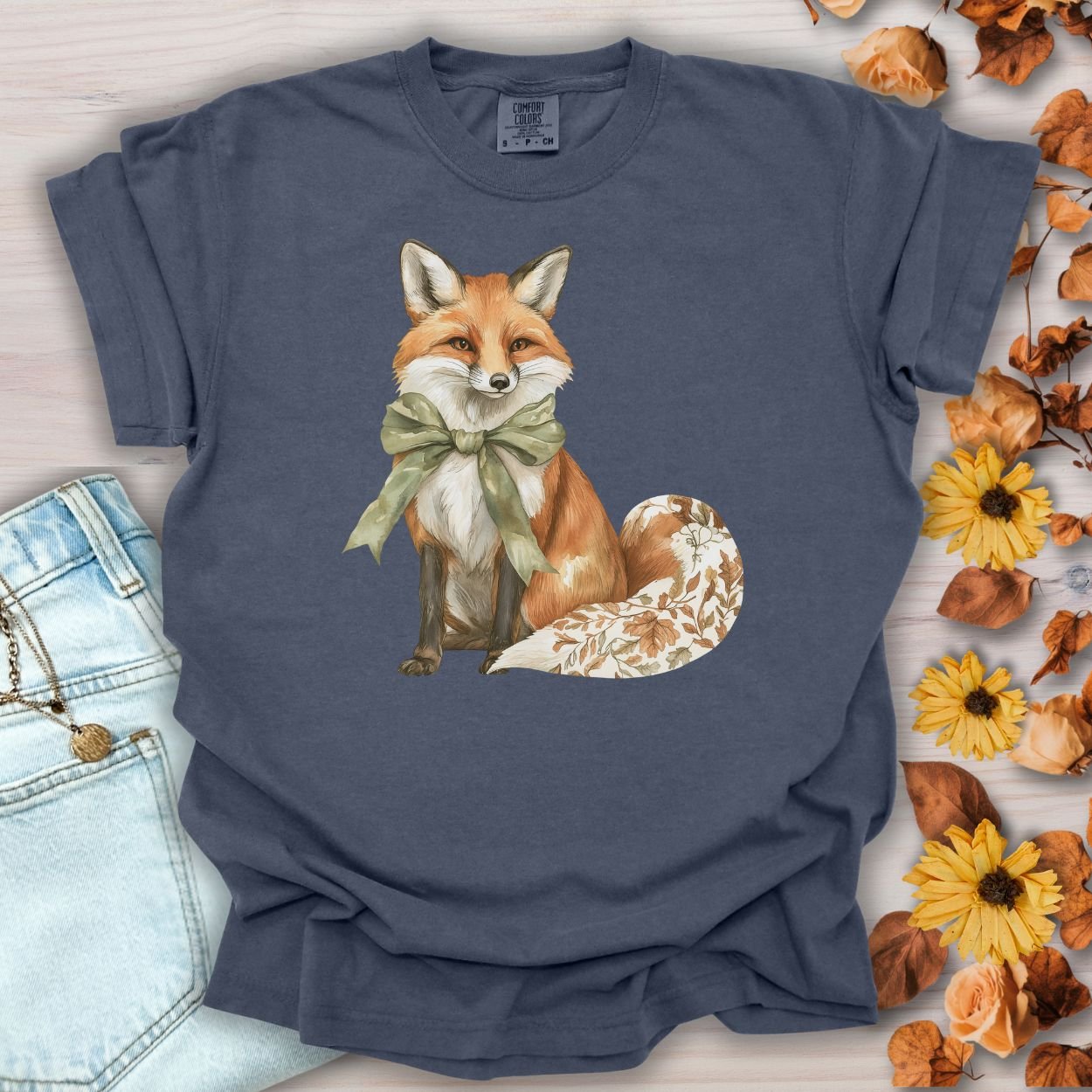 Denim blue garment-dyed t-shirt with a watercolor fox wearing a sage-green bow and leaf-patterned tail.
