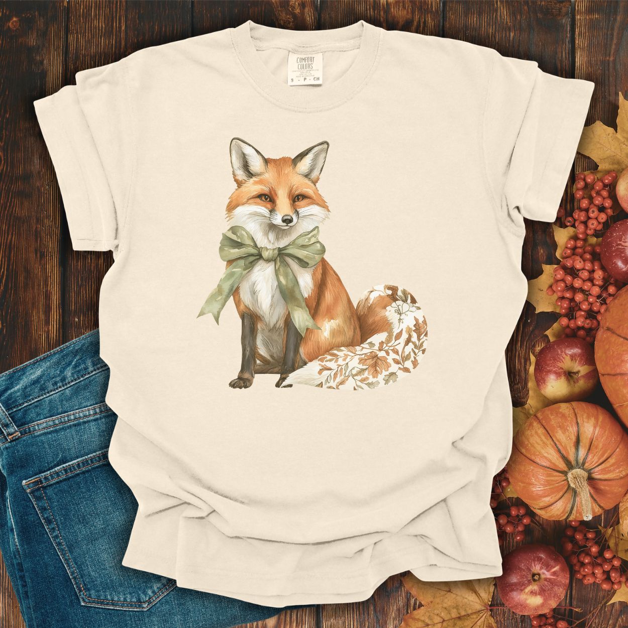 Ivory garment-dyed t-shirt with a watercolor fox wearing a sage-green bow and a leaf-patterned tail.